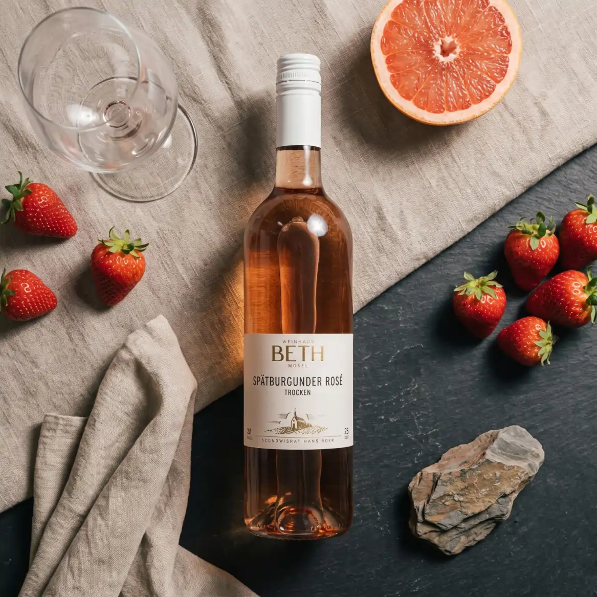 Eine Flasche Rosé vom Weinhaus Beth sommerlich liegend drapiert auf deinem Tisch mit Grapefruit und Erdbeeren